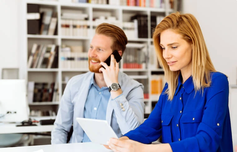 Business professionals collaborating in an office, with a woman reviewing documents and a man talking on the phone.