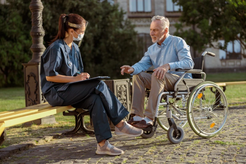 Healthcare professional consulting an elderly man in a wheelchair outdoors.